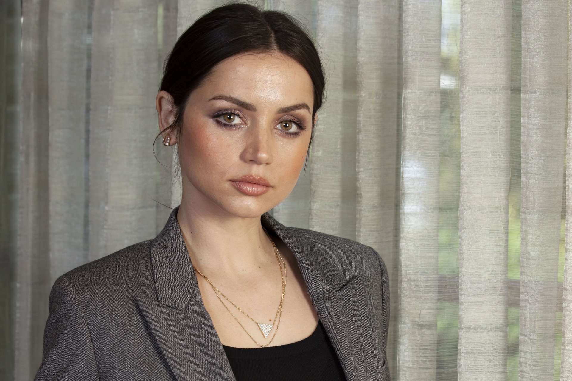 Close-up of Cuban actress Ana de Armas with green eyes and brunette hair, wearing a gray jacket against a softly lit curtain background.