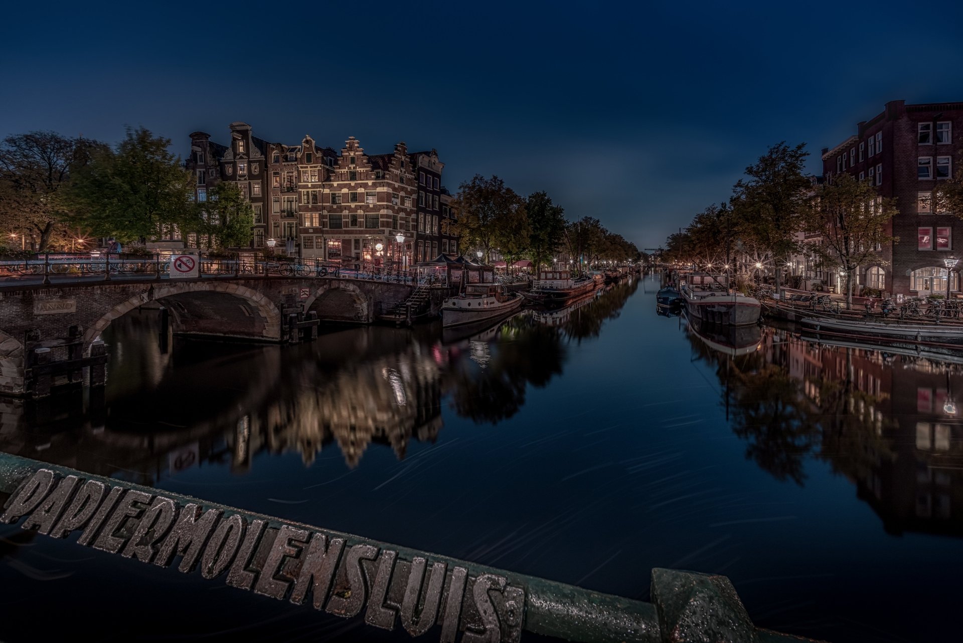 Night view of an Amsterdam canal in the Netherlands with historic houses and buildings reflected in calm water, illuminated man-made bridges and moored boats — 5K Ultra HD PC desktop wallpaper.
