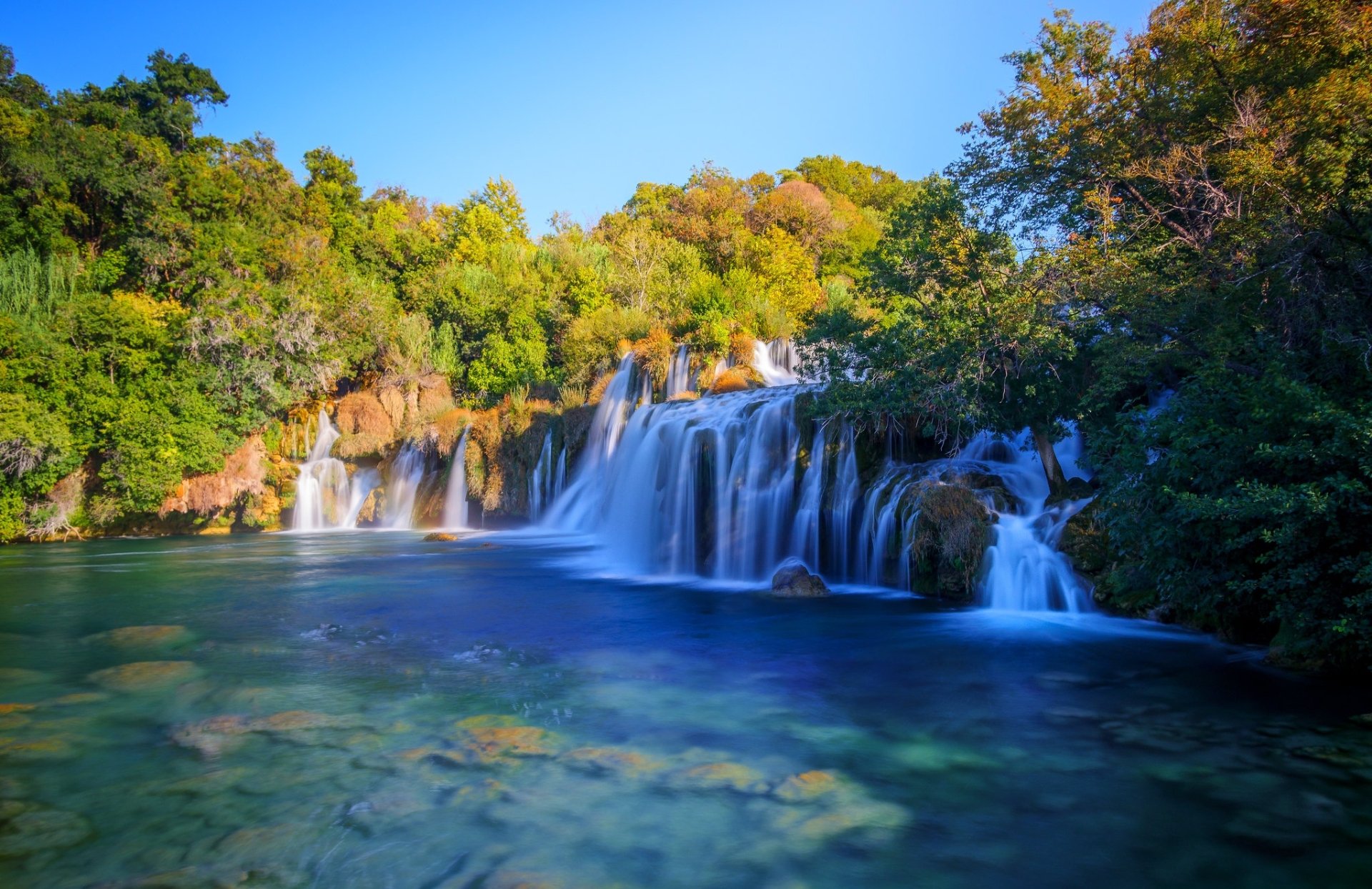 HD desktop wallpaper showcasing a stunning waterfall in Croatia surrounded by lush green trees under a clear blue sky.