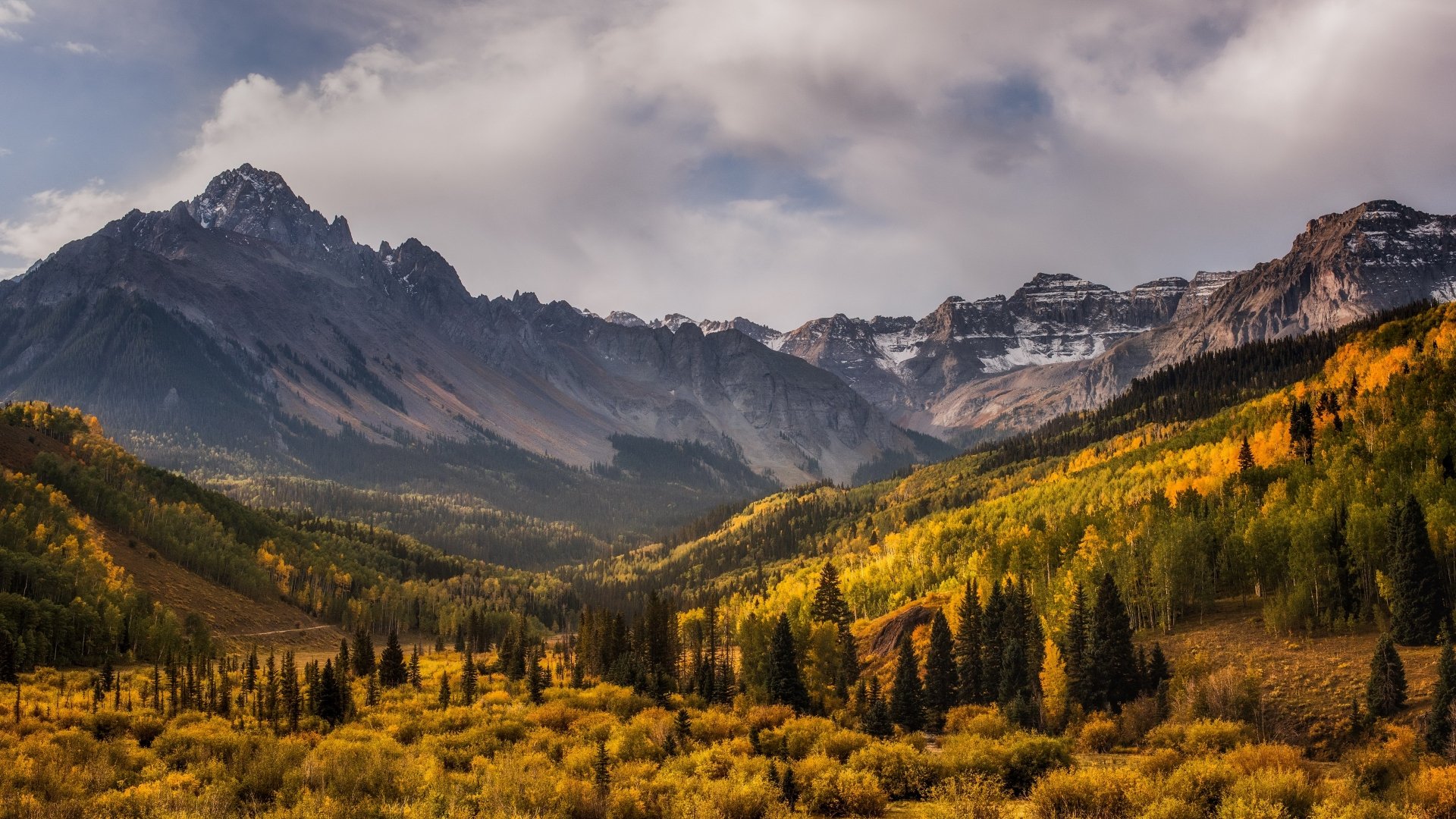 4K Ultra HD PC desktop wallpaper — fall mountain landscape: golden aspen forest filling a valley beneath jagged peaks and a dramatic sky.