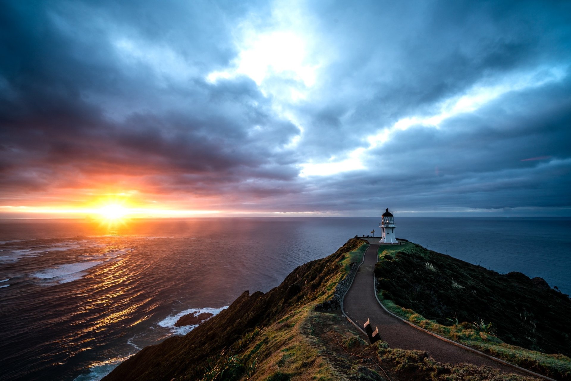 HD PC wallpaper: lighthouse atop a rugged New Zealand headland at sunset, winding road to the tower, Pacific Ocean and dramatic cloudy sky.