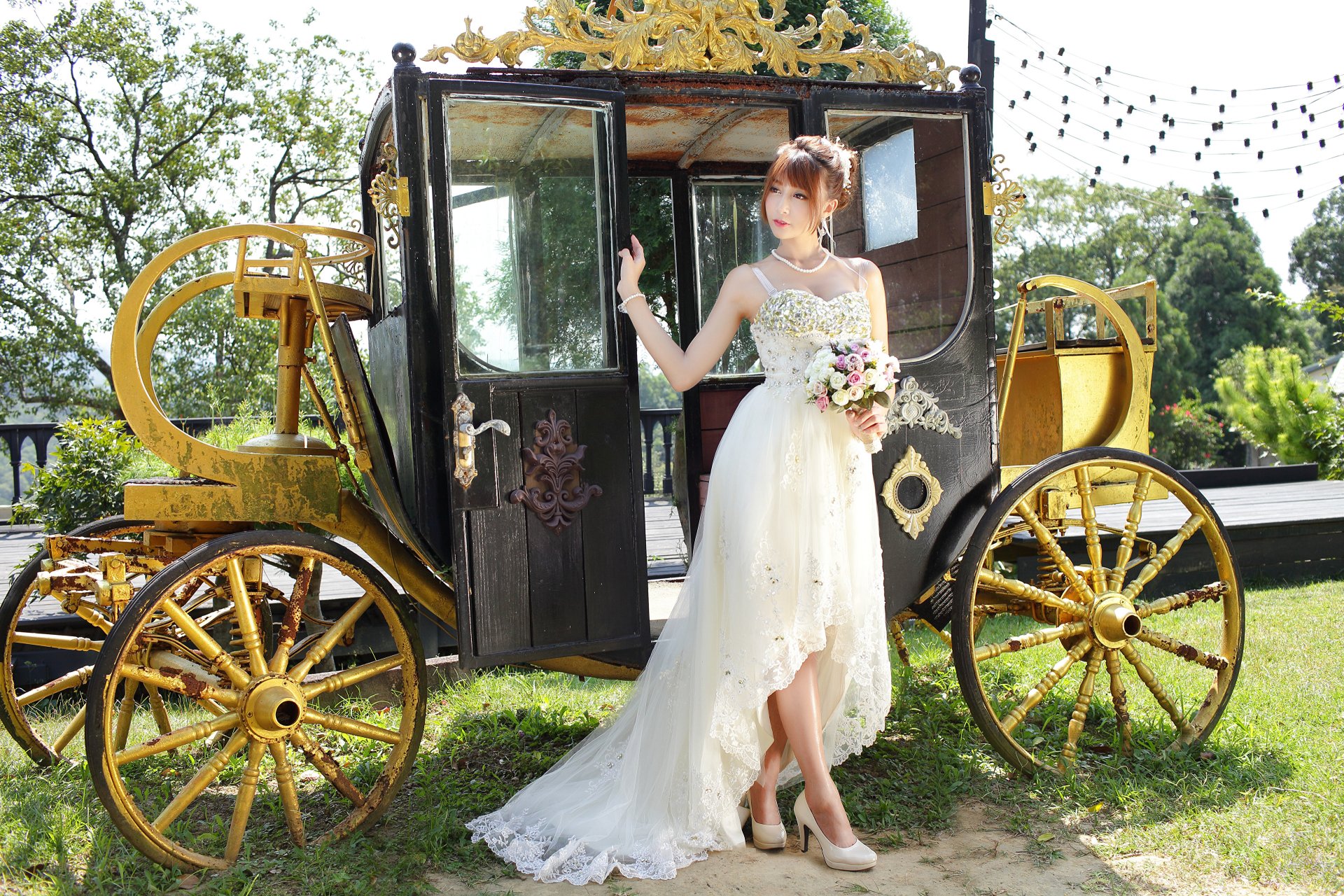 An Asian brunette bride in a white wedding dress poses gracefully beside an ornate golden carriage in a lush outdoor setting, captured in 4K Ultra HD.