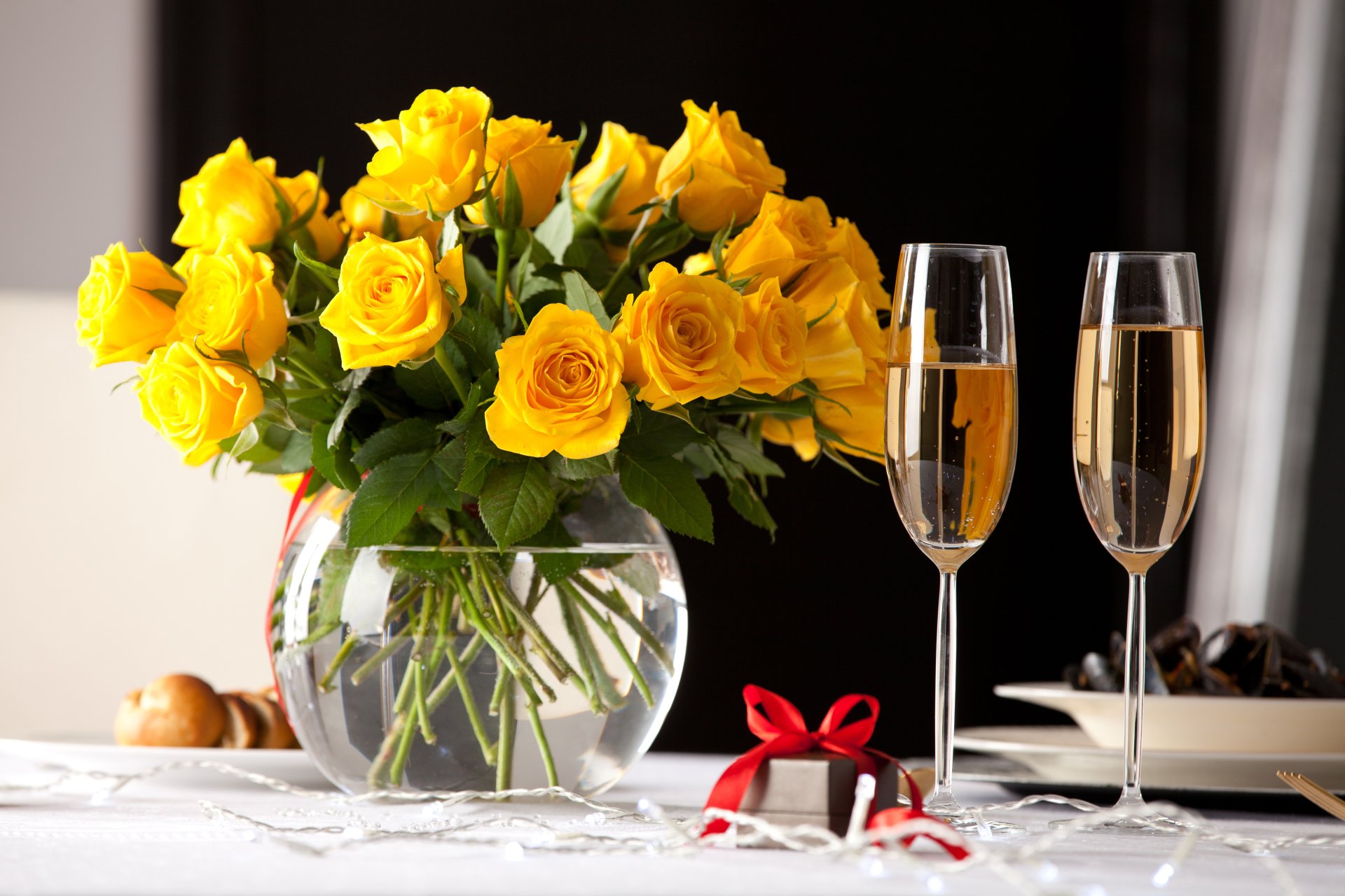 A 4K Ultra HD still life desktop wallpaper featuring a vase of vibrant yellow roses, two champagne glasses, and a small red gift box on a white table.