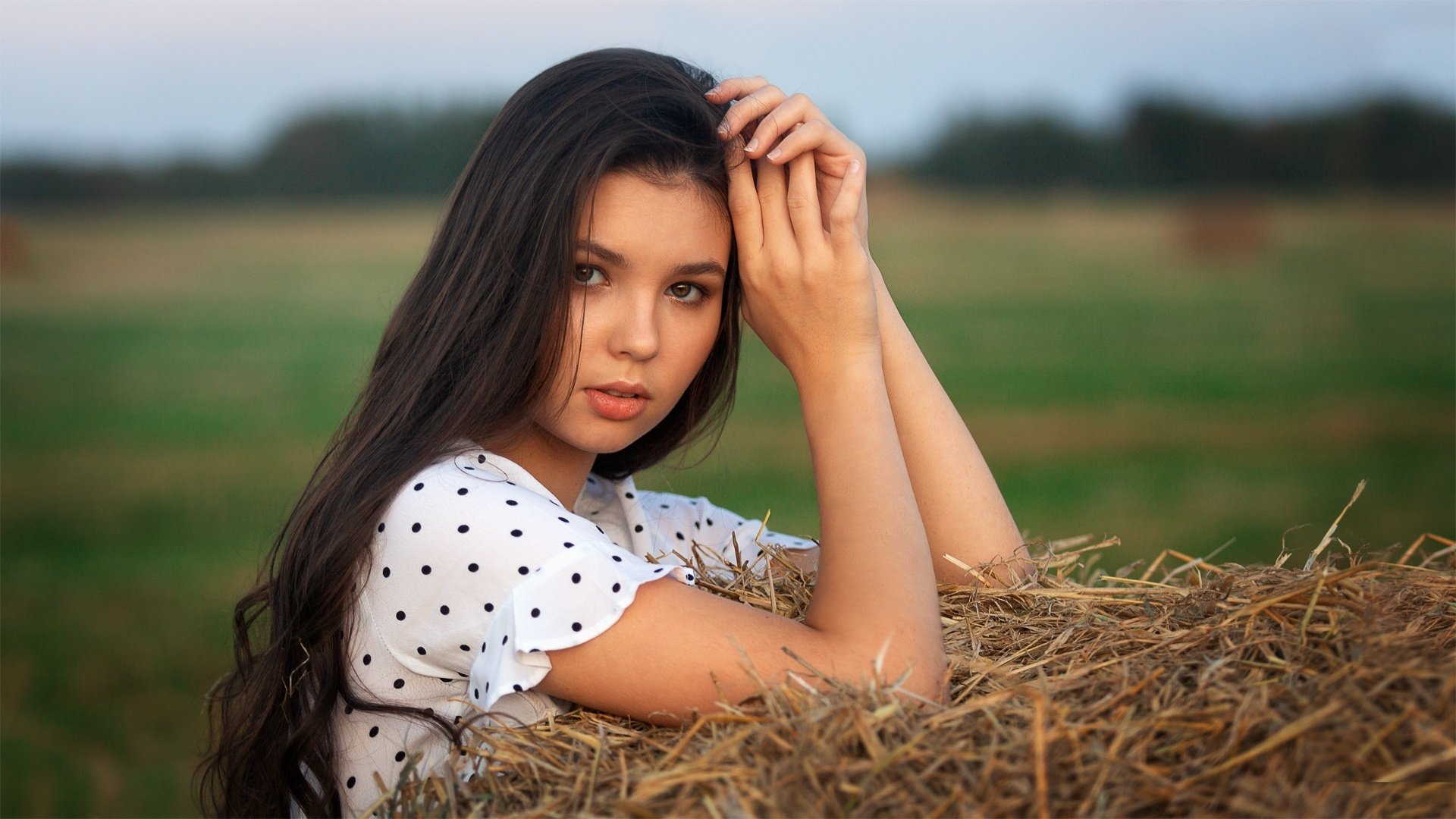 HD PC desktop wallpaper of a long-haired woman model with black hair leaning on a haystack, soft rural background and shallow depth of field.