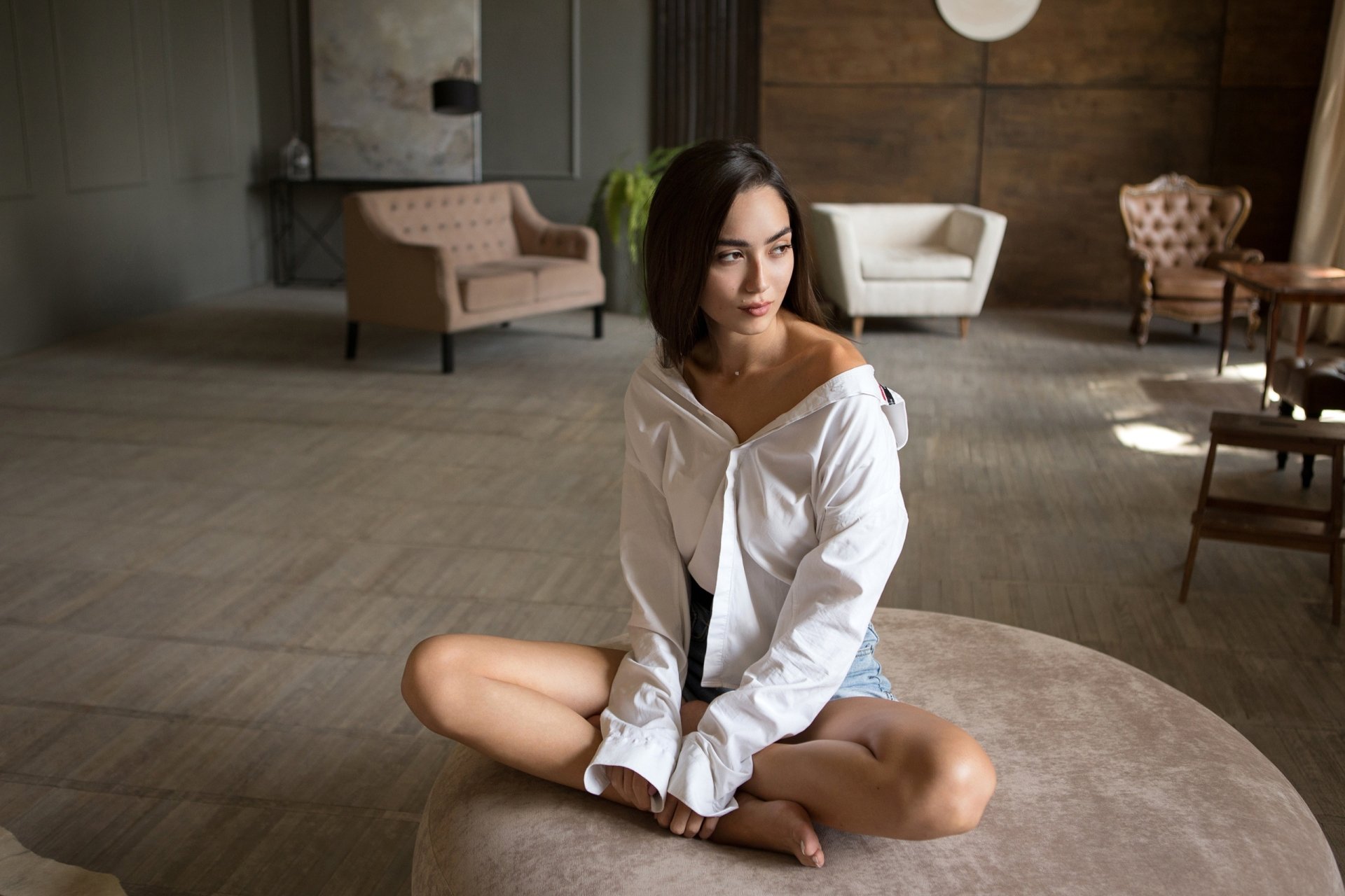 Brunette model in shorts sits cross-legged on a round surface in a softly lit, stylish living room, creating a calm and introspective mood. HD PC desktop wallpaper.