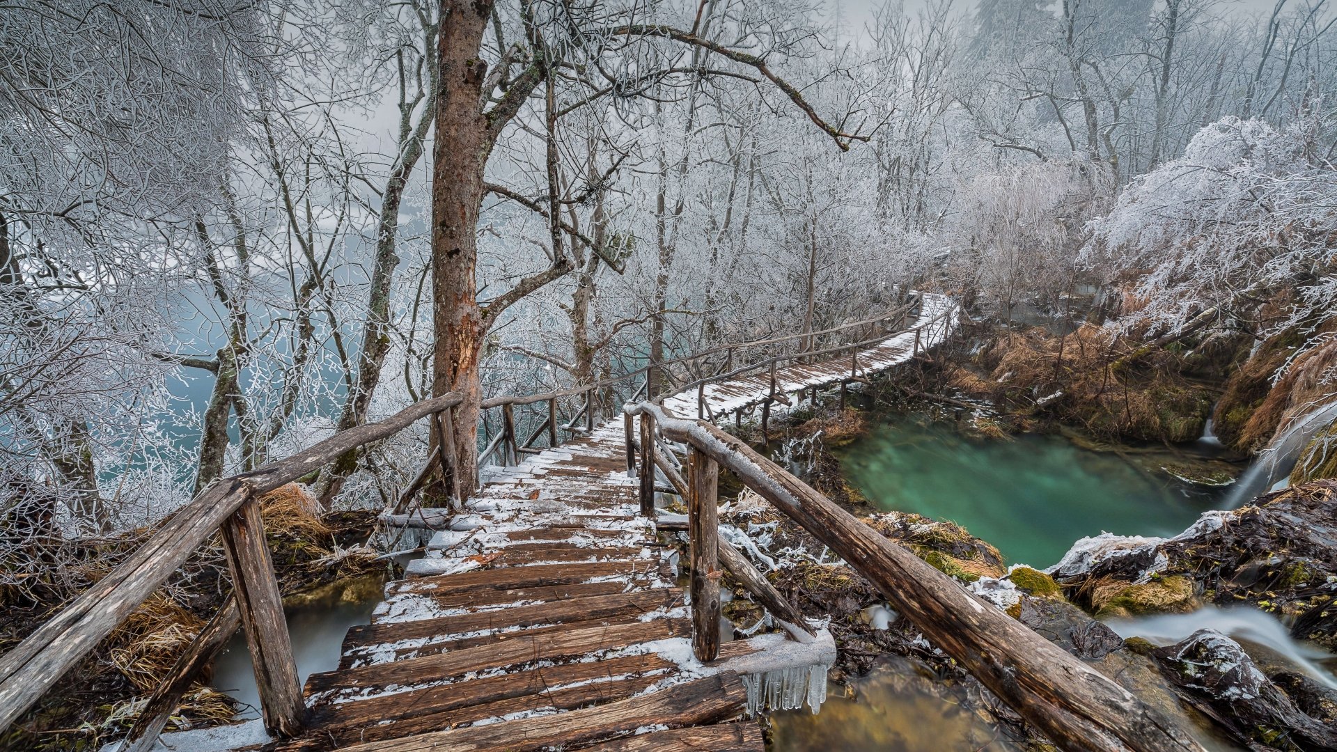 Winter scene at Plitvice Lakes National Park, Croatia, showing a wooden bridge over turquoise water surrounded by frost-covered trees. 4K Ultra HD desktop wallpaper.