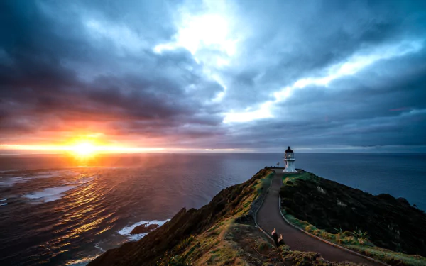 HD PC wallpaper: lighthouse atop a rugged New Zealand headland at sunset, winding road to the tower, Pacific Ocean and dramatic cloudy sky.