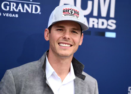 HD wallpaper of a smiling man in a baseball cap and a grey blazer posing for a photo.