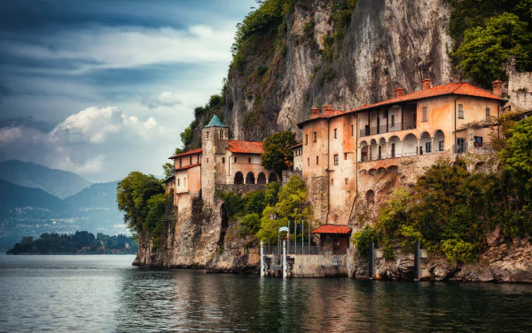 HD PC desktop wallpaper: historic religious monastery clinging to a cliff above Lake Maggiore, Lombardy, Italy, with calm water and dramatic skies.