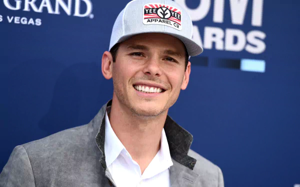 HD wallpaper of a smiling man in a baseball cap and a grey blazer posing for a photo.
