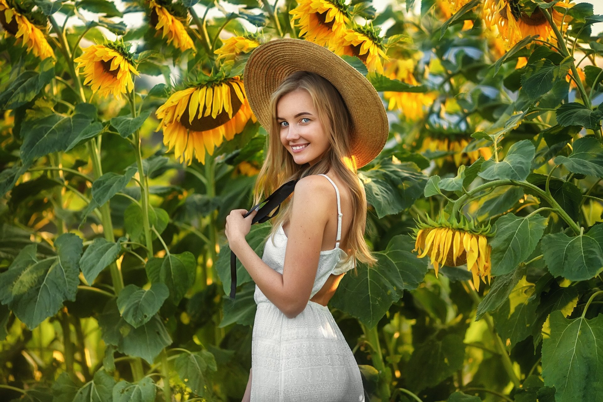 Blonde woman in a white dress and wide-brimmed hat smiles while standing in a vibrant sunflower field, captured in HD for a desktop wallpaper background.