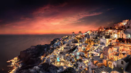 Night view of Santorini, Greece, showcasing illuminated white buildings cascading down the cliffs under a vibrant, colorful sky, captured in HD for a desktop background.