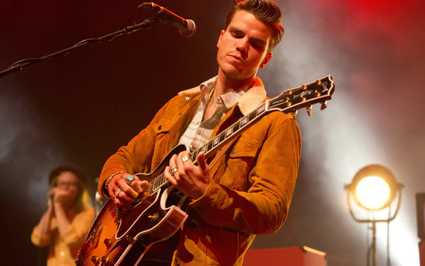 HD wallpaper featuring KALEO's Jökull Júlíusson playing guitar on stage with warm stage lighting in the background.