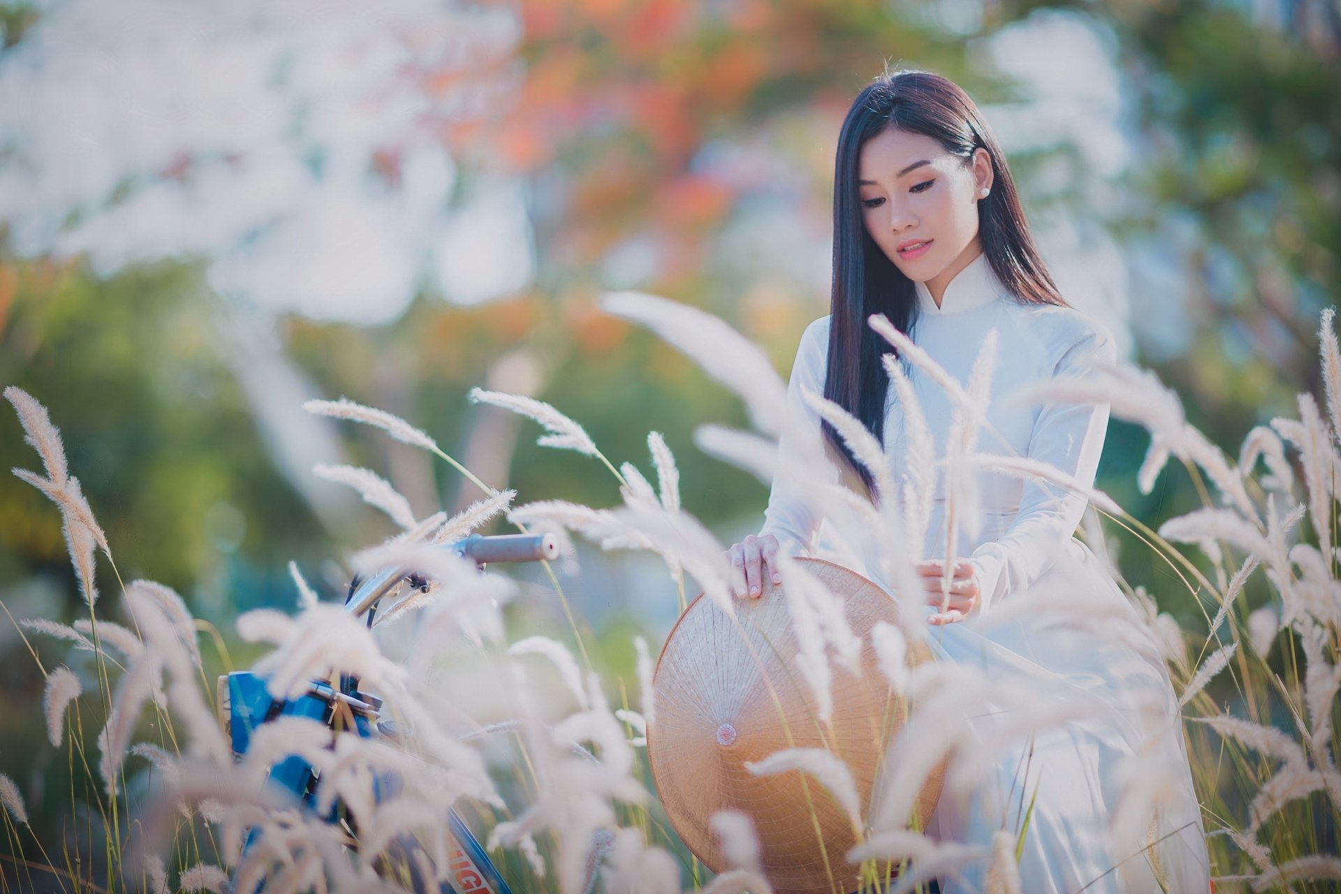 A brunette Asian woman wearing a white Ao Dai and traditional hat poses amidst tall grasses with a soft depth of field in this 4K Ultra HD desktop wallpaper.
