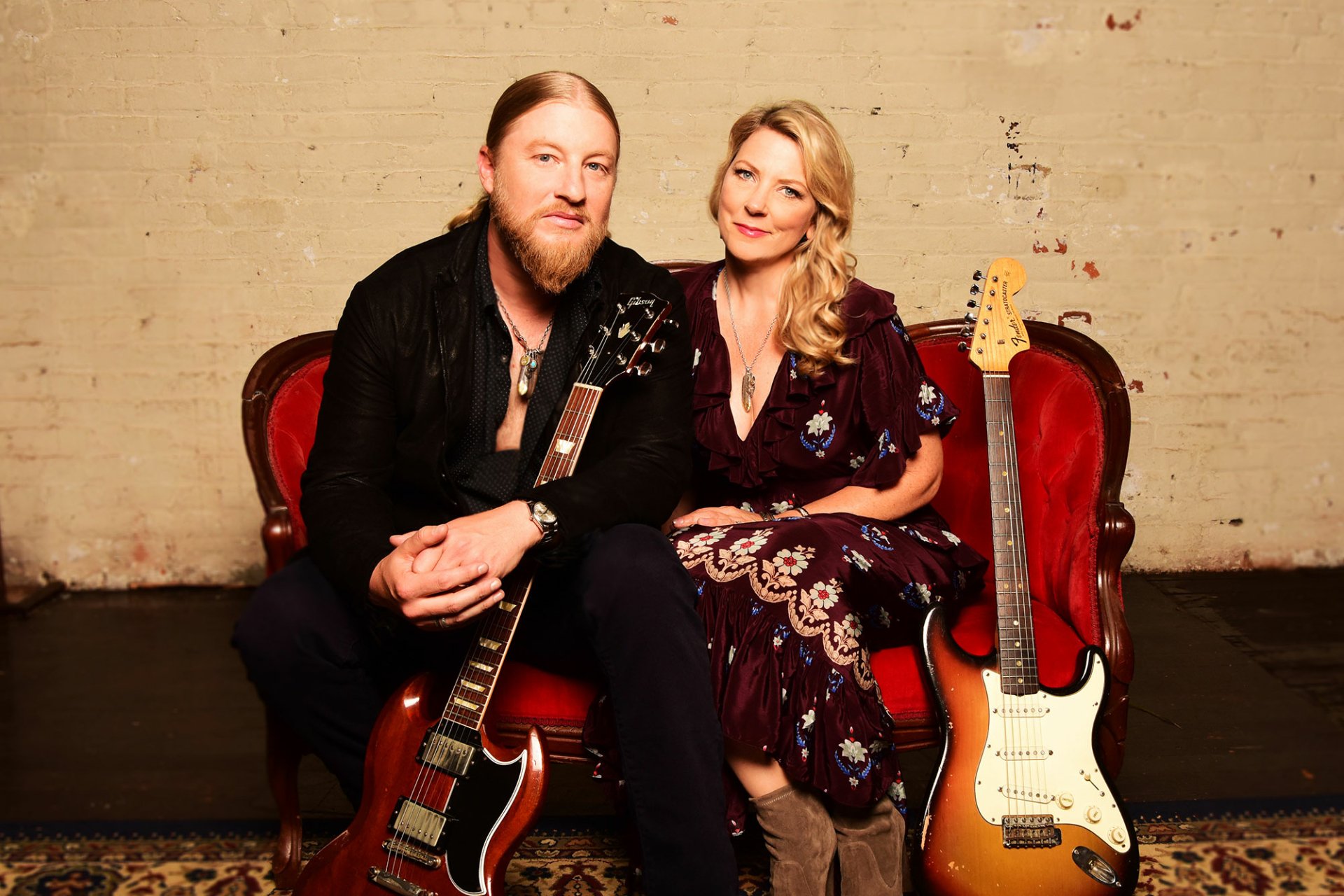 HD wallpaper of Tedeschi Trucks Band with a man holding a guitar and a woman beside him, both seated on red chairs.