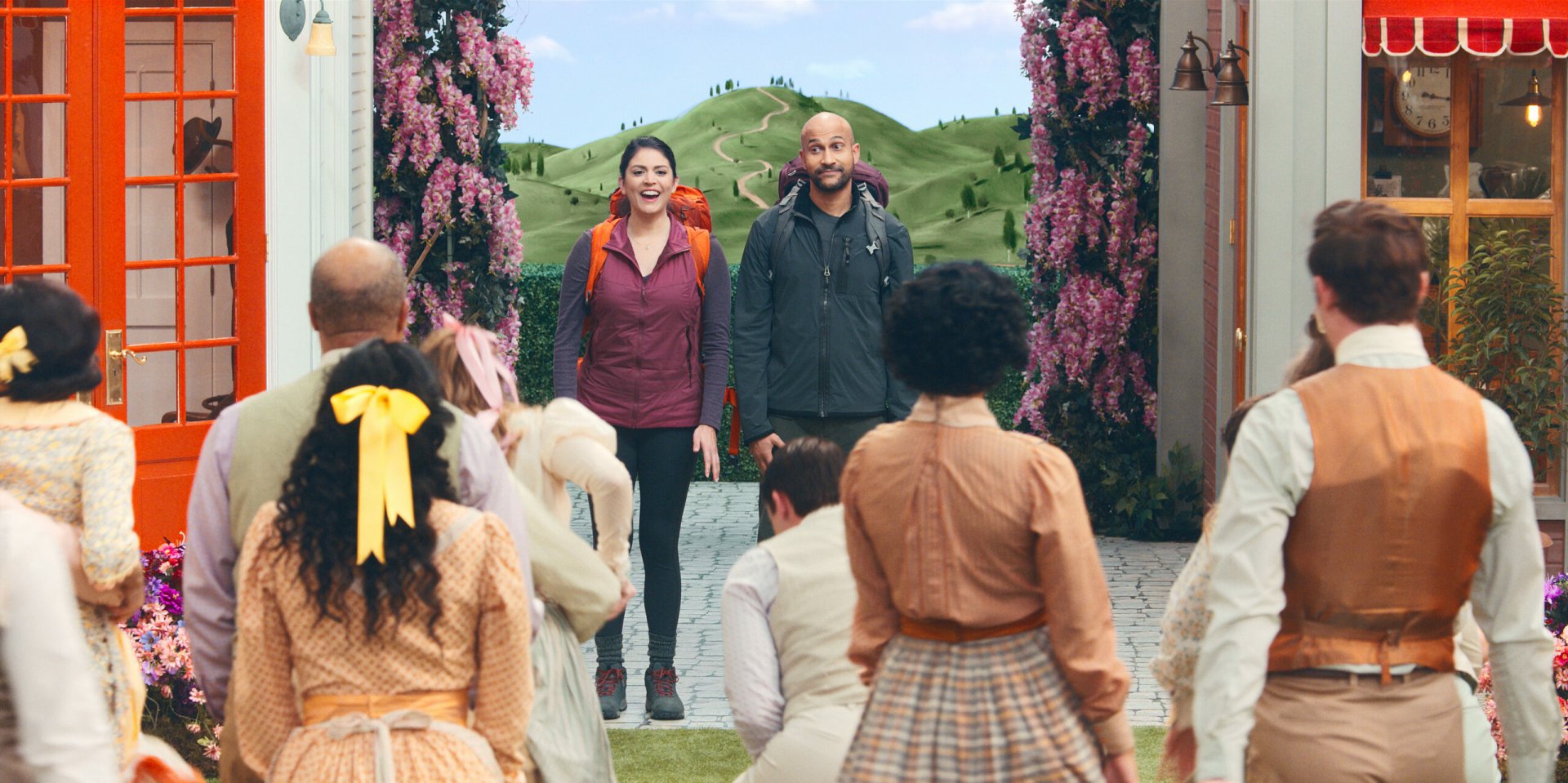 HD desktop wallpaper featuring a scene from Schmigadoon! with a man and a woman entering a vibrant, vintage-style town as onlookers gaze towards them, amidst lush green hills and blooming flowers.