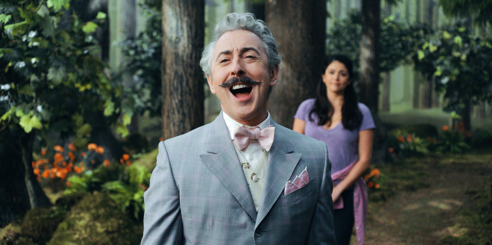 HD wallpaper featuring a cheerful man in a grey suit with a bow tie and a woman in purple standing in a forest from Schmigadoon!
