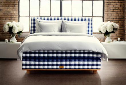A stylish bed with a blue and white checkered pattern, complemented by soft white bedding, set against a backdrop of large windows and elegant floral arrangements.