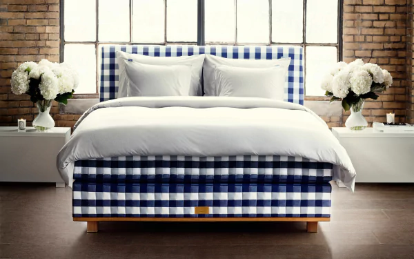 A stylish bed with a blue and white checkered pattern, complemented by soft white bedding, set against a backdrop of large windows and elegant floral arrangements.