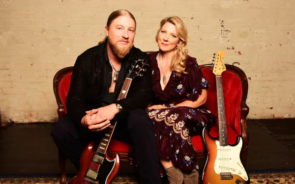 HD wallpaper of Tedeschi Trucks Band with a man holding a guitar and a woman beside him, both seated on red chairs.