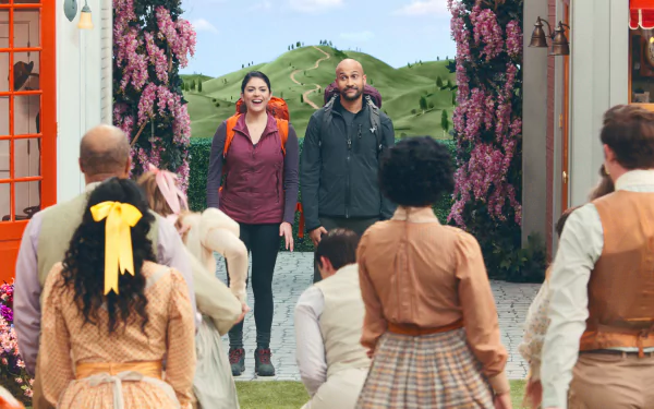 HD desktop wallpaper featuring a scene from Schmigadoon! with a man and a woman entering a vibrant, vintage-style town as onlookers gaze towards them, amidst lush green hills and blooming flowers.