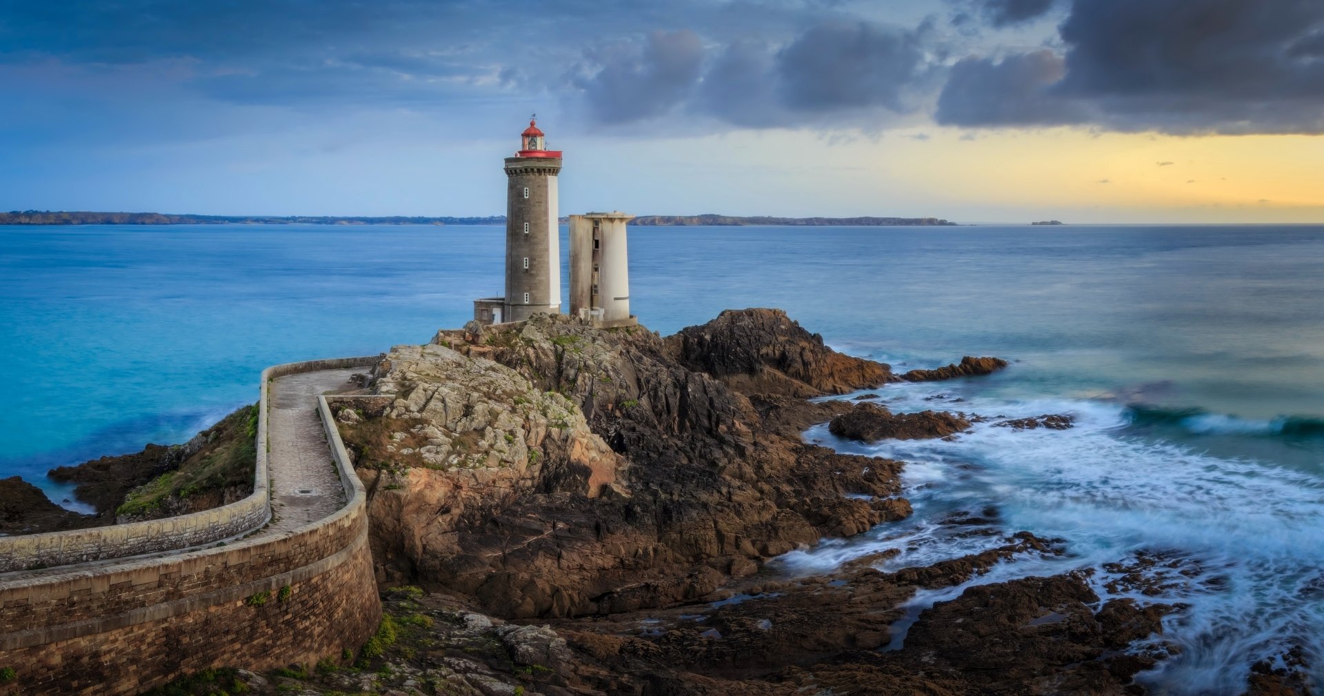 5K Ultra HD PC desktop wallpaper/background: man-made lighthouse on a rocky promontory with stone causeway, churning sea and dramatic evening sky.