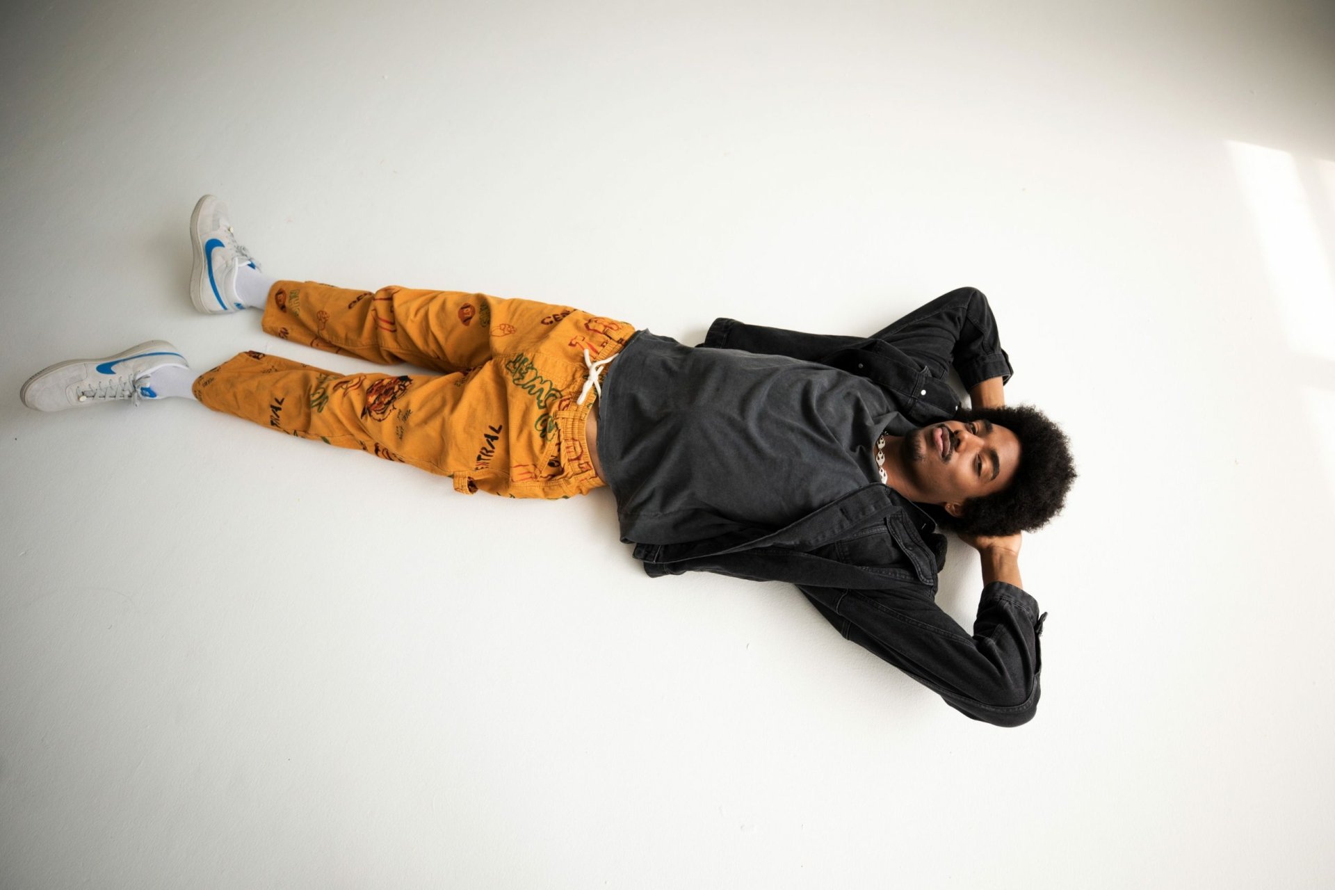 A person relaxing on the floor with a casual pose, wearing orange pants and a grey jacket, against a white background - HD Tai Verdes-themed desktop wallpaper.