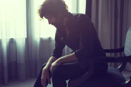 Stylish HD wallpaper featuring a thoughtful man sitting on a chair against a softly lit curtain backdrop.