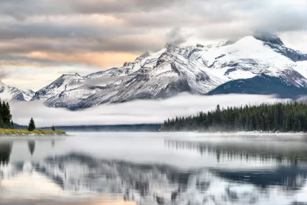 A serene 4K Ultra HD desktop wallpaper of Maligne Lake, showcasing snow-capped mountains reflected on the calm water surrounded by mist and dense forest under a cloudy sky.