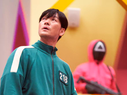 2K Quad HD Squid Game TV wallpaper: contestant in green tracksuit numbered 218 looks upward as a masked guard with triangle symbol stands behind in a bright game hall.