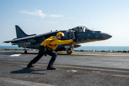 HD desktop wallpaper featuring a McDonnell Douglas AV-8B Harrier II jet fighter on a carrier deck with a crew member signaling in the foreground.