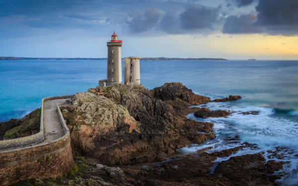 5K Ultra HD PC desktop wallpaper/background: man-made lighthouse on a rocky promontory with stone causeway, churning sea and dramatic evening sky.