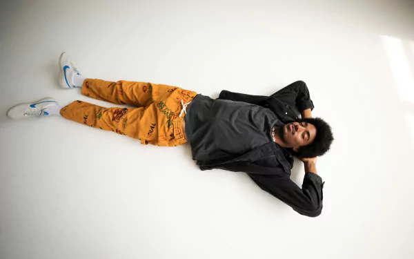 A person relaxing on the floor with a casual pose, wearing orange pants and a grey jacket, against a white background - HD Tai Verdes-themed desktop wallpaper.