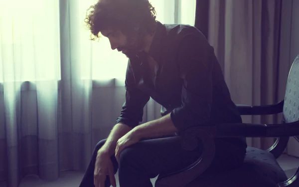 Stylish HD wallpaper featuring a thoughtful man sitting on a chair against a softly lit curtain backdrop.