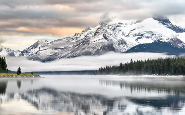 A serene 4K Ultra HD desktop wallpaper of Maligne Lake, showcasing snow-capped mountains reflected on the calm water surrounded by mist and dense forest under a cloudy sky.