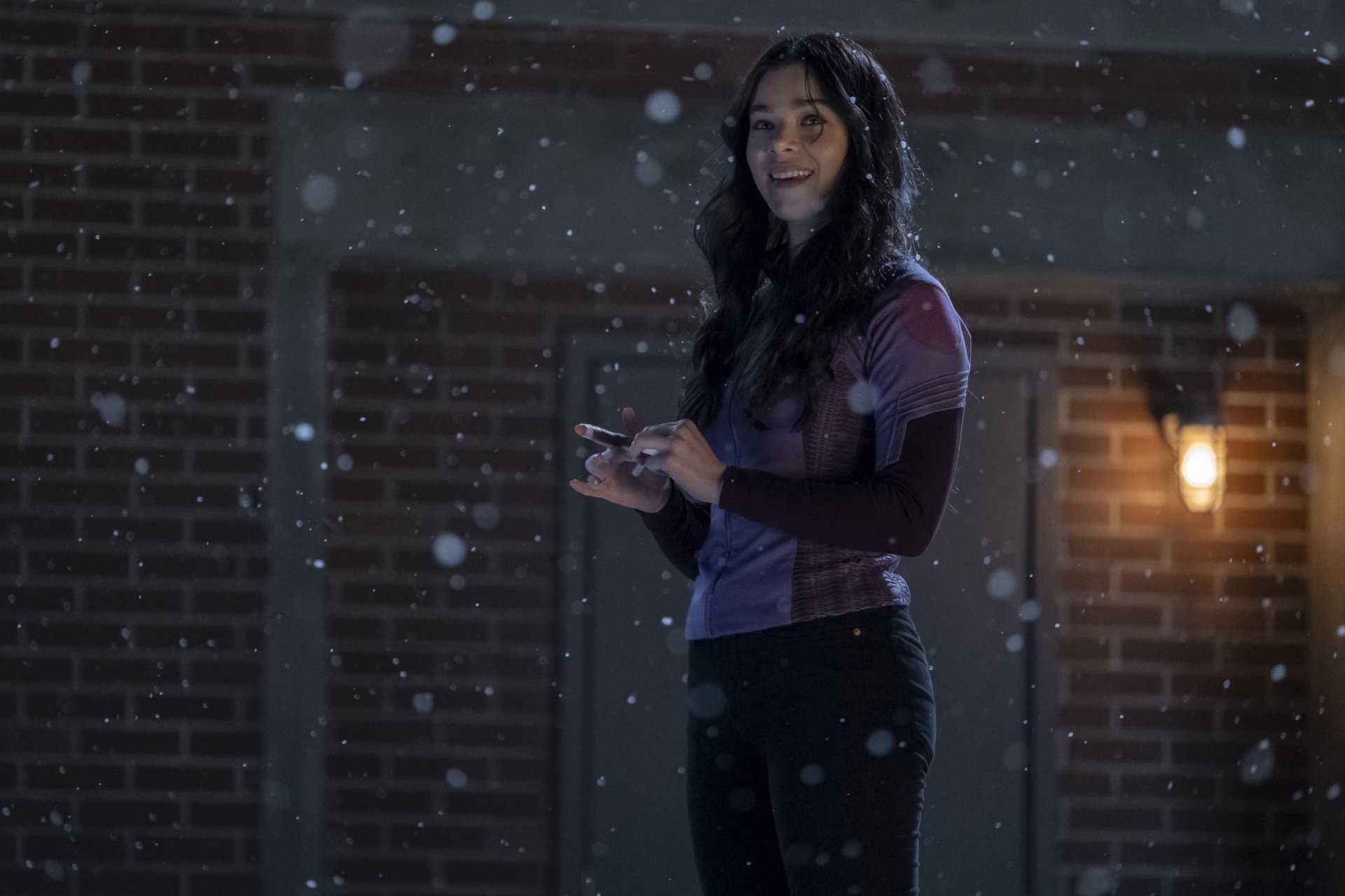 2K Quad HD PC wallpaper: young woman in a purple top standing in falling snow on a dim brick street, giving a TV-show archer vibe.