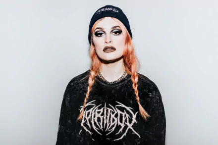 A striking HD desktop wallpaper featuring a musician wearing a Spiritbox shirt and beanie, with vibrant orange hair and bold makeup, conveying a powerful music vibe.