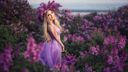HD PC desktop wallpaper: blonde model in a lilac dress and floral wreath standing amid purple lilac blossoms with a soft coastal horizon background.