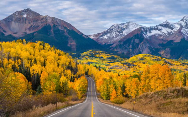4K Ultra HD desktop wallpaper featuring a winding road cutting through a vibrant fall landscape with golden trees and towering mountains under a partly cloudy sky.