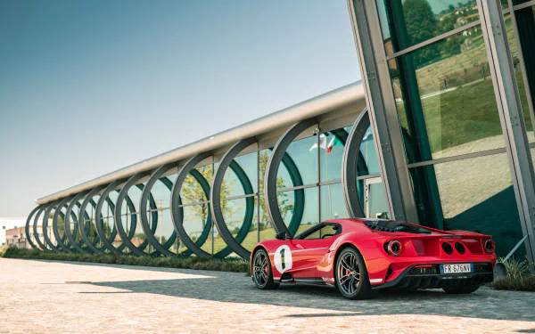 Red Ford GT parked beside a modern glass building under clear blue sky, captured in stunning 4K Ultra HD for PC desktop wallpaper and background.