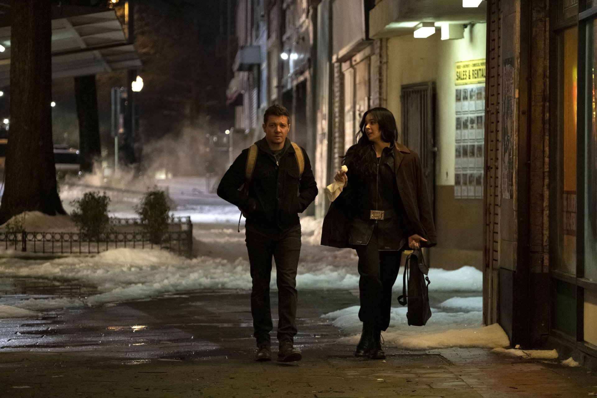 Hailee Steinfeld as Kate Bishop and Jeremy Renner as Clint Barton walk together in a snowy urban street scene from the TV show Hawkeye, featured in an HD desktop wallpaper.