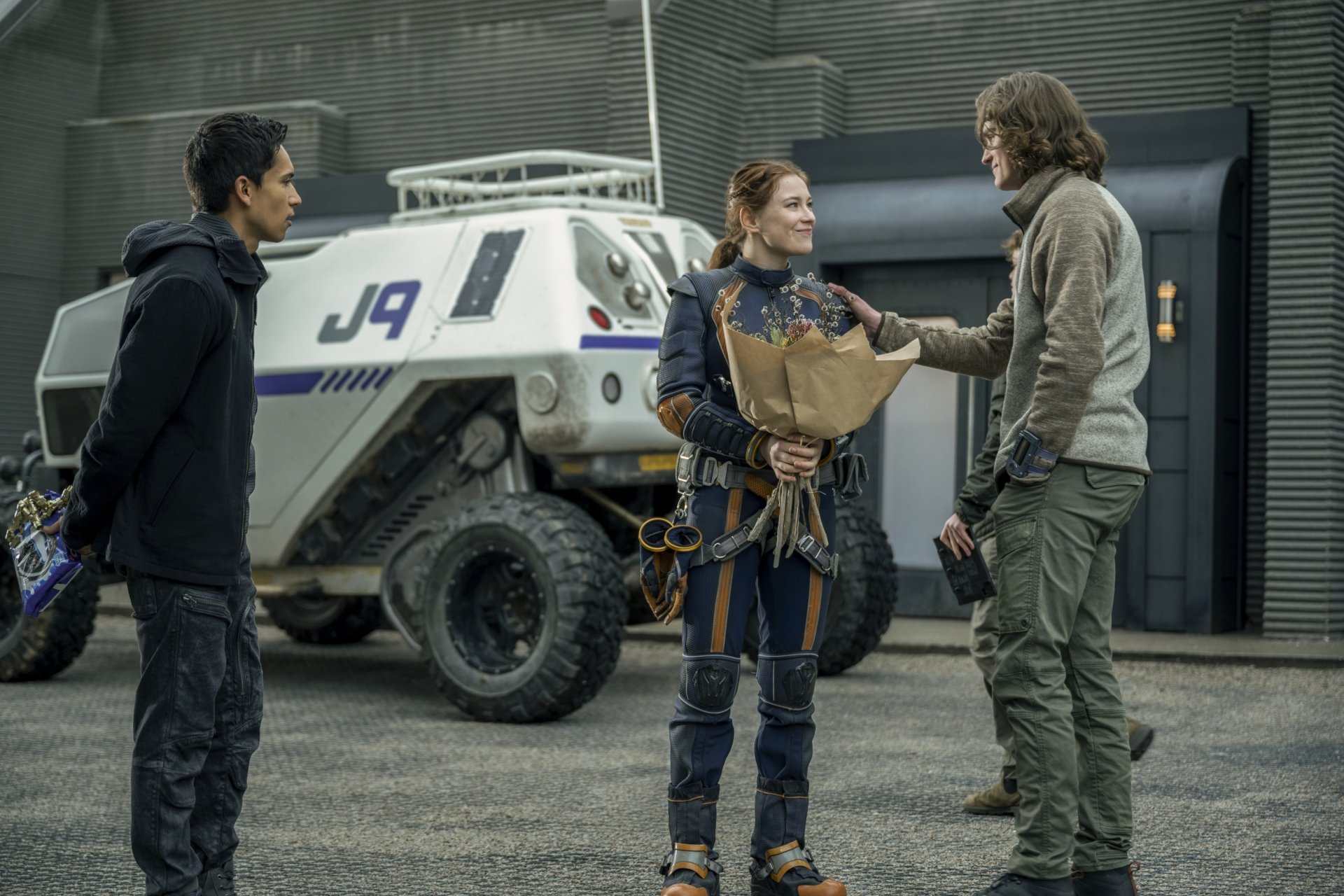 2K Quad HD PC wallpaper: three Lost In Space characters stand by a J9 rover in a metallic hangar; one holds a bouquet as the others converse.