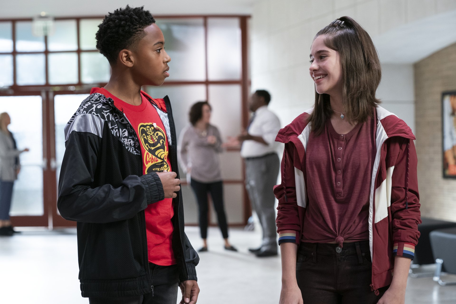 Cobra Kai TV show 2K Quad HD PC desktop wallpaper: two high school students—a teen boy in a red shirt and jacket and a smiling teen girl in a maroon hoodie—talking in a school hallway.