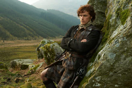 Jamie Fraser from the TV show Outlander, in Highland dress leaning against a mossy rock amid misty glens — 5K Ultra HD PC desktop wallpaper.