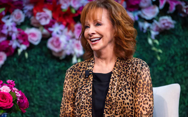 HD desktop wallpaper featuring a smiling woman with red hair, wearing a leopard print jacket, seated in front of a floral background.