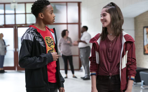Cobra Kai TV show 2K Quad HD PC desktop wallpaper: two high school students—a teen boy in a red shirt and jacket and a smiling teen girl in a maroon hoodie—talking in a school hallway.