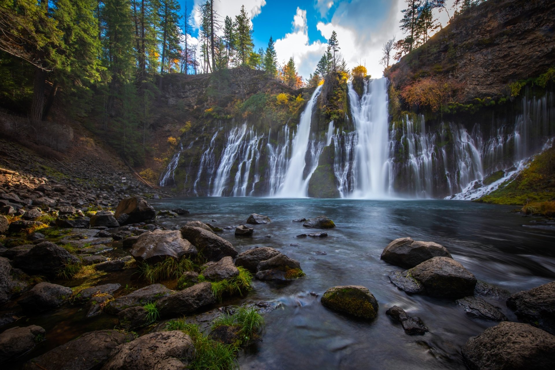 HD PC desktop wallpaper of Burney Falls: multi-tiered waterfall plunging into a clear pool, mossy rocks in foreground and pine trees framing a bright sky — nature background.