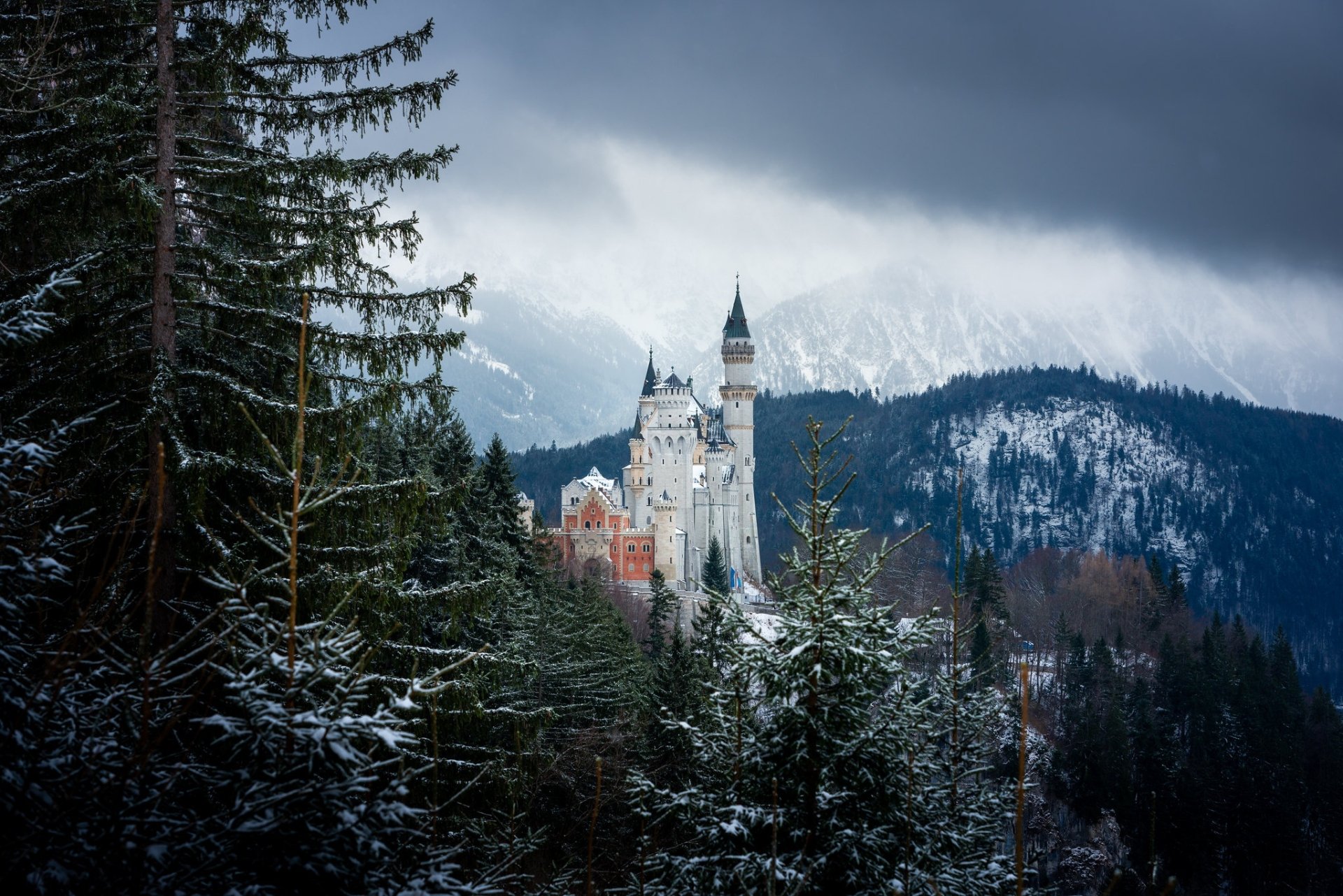 Neuschwanstein Castle stands majestically amidst snowy pine trees in the Bavarian Alps, Germany, under a moody, overcast sky.