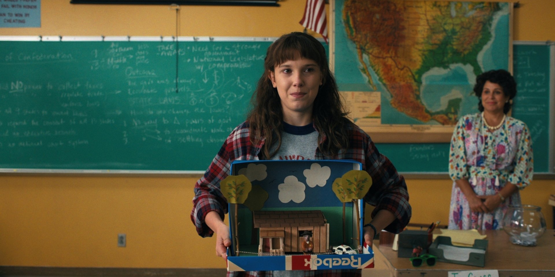 Eleven from Stranger Things, played by Millie Bobby Brown, stands in a classroom holding a model house, featured in this HD PC desktop wallpaper background.