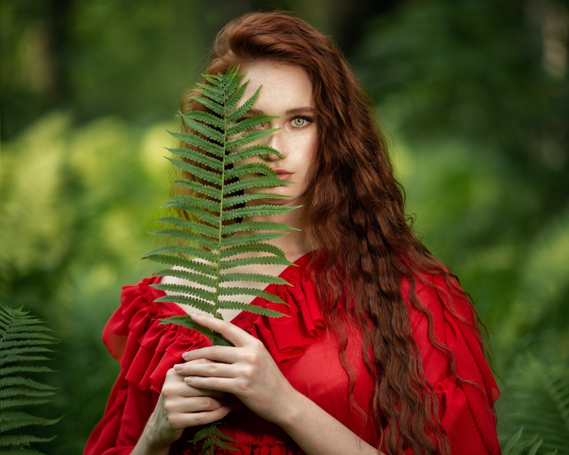 HD desktop wallpaper showing a redhead woman model with long curly hair holding a fern leaf partially covering her face against a blurred green background.